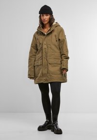 Parka verde oliva con cappuccio, bottoni frontali, due grandi tasche a toppa e dettagli con cordino. Indossata sopra leggings neri e stivali chunky.