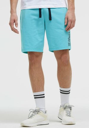 Männlich, trägt hellblaue Freizeithosen, weiße Socken mit zwei schwarzen Streifen und weiße Turnschuhe, steht vor einem schlichten Hintergrund.
