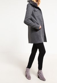 Cappotto grigio in misto lana con cappuccio in pelliccia sintetica, dotato di due tasche frontali e chiusura con bottone, abbinato a leggings neri e stivaletti grigi. 