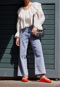 Kvinde iført hvid broderet bluse, vidde blå jeans, klare orange sandaler og bærende på en lille sort vævet håndtaske udendørs.