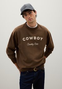 Felpa marrone con ricamo in off-white "COWBOY" e "Country Club"; abbinata a jeans blu scuro e un cappellino grigio con logo 'P'.