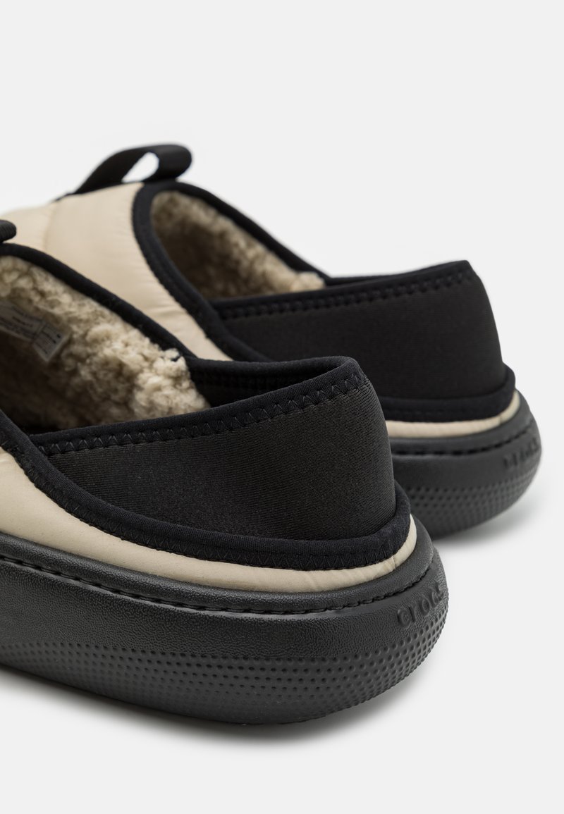 Zapatos tipo slip-on con parte superior negra y acentos beige, forro de felpa suave y suela de goma texturizada para agarre y comodidad.