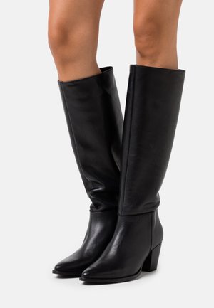 Bottes en cuir noir montantes jusqu'aux genoux avec talons blocs portées sur des jambes nues sur un fond clair uni.