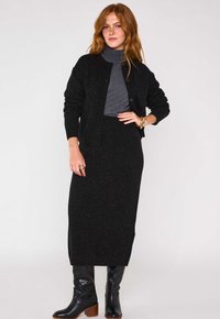 Cardigan nero con scollatura rotonda e bottoni frontali, indossato sopra un dolcevita grigio scuro. Una lunga gonna nera e stivali neri fino al ginocchio completano l'outfit.