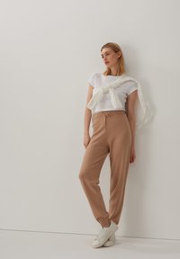 Leichtes weißes T-Shirt, bequeme beige Jogginghose mit Kordelzug und weiße Sneaker. Die Hose hat eine lockere Passform; das T-Shirt ist lässig und figurbetont.