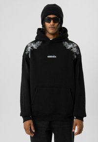 Zwarte hoodie met bloemenborduursel op de schouders, voorzak en witte tekst "PASSIE VOOR HET LEVEN" op de borst. G gedragen met een zwarte beanie en zonnebril.