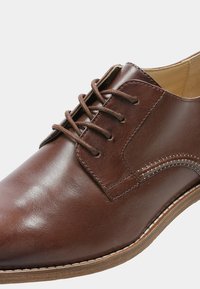 Chaussure habillée en cuir marron avec une finition lisse, dotée de lacets ronds, d'accents cousus et d'une semelle beige clair. Design classique à bout rond.