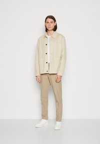 Veste en coton beige avec fermeture à boutons et poche poitrine, associée à un pantalon marron clair et des sneakers blanches. Design simple et minimal.