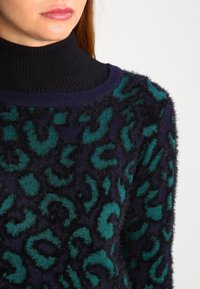 Maglione navy fuzzy con stampa leopardata turchese, caratterizzato da un collo alto nero a costine. La consistenza morbida aggiunge calore e profondità al design.