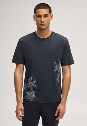 Man wearing a black short-sleeve t-shirt with subtle palm tree prints on the lower front and black pants, standing against a plain background.
