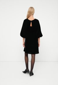 Rochie din catifea neagră cu detaliu în formă de cheiță la spate și mâneci late tip clopot. Rochia se termină deasupra genunchilor, fiind complementată de colanți negri transparenți și pantofi.