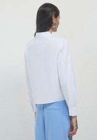 Chemise blanche à manches longues avec col, dotée d'un ourlet droit et de poignets à boutons, associée à un pantalon bleu clair. Texture lisse, design minimal.