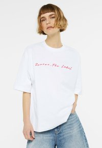 White oversized t-shirt made of cotton, featuring red cursive text "Senses. The Label" on the front, with short sleeves and a relaxed fit.