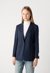 Navy pinstripe blazer with a tailored fit, single button closure, notched lapels, and side pockets, paired with a white turtleneck.