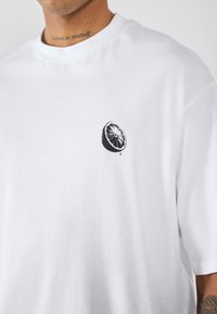 Hvid kortærmet t-shirt i et blødt stof med en sort citrusfrugt-illustration på venstre brystområde. Glat tekstur.