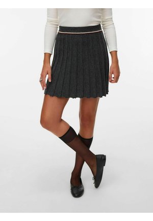 Femme portant une jupe plissée gris foncé, des chaussettes noires transparentes montant jusqu'aux genoux, des ballerines noires et un haut blanc à manches longues, debout avec les jambes croisées.