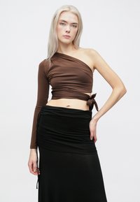 Smeđi asimetrični kratki top s dugim rukavima i detaljem vezanja na struku, u kombinaciji s dugom crnom suknjom s naborima. Glatka tekstura.