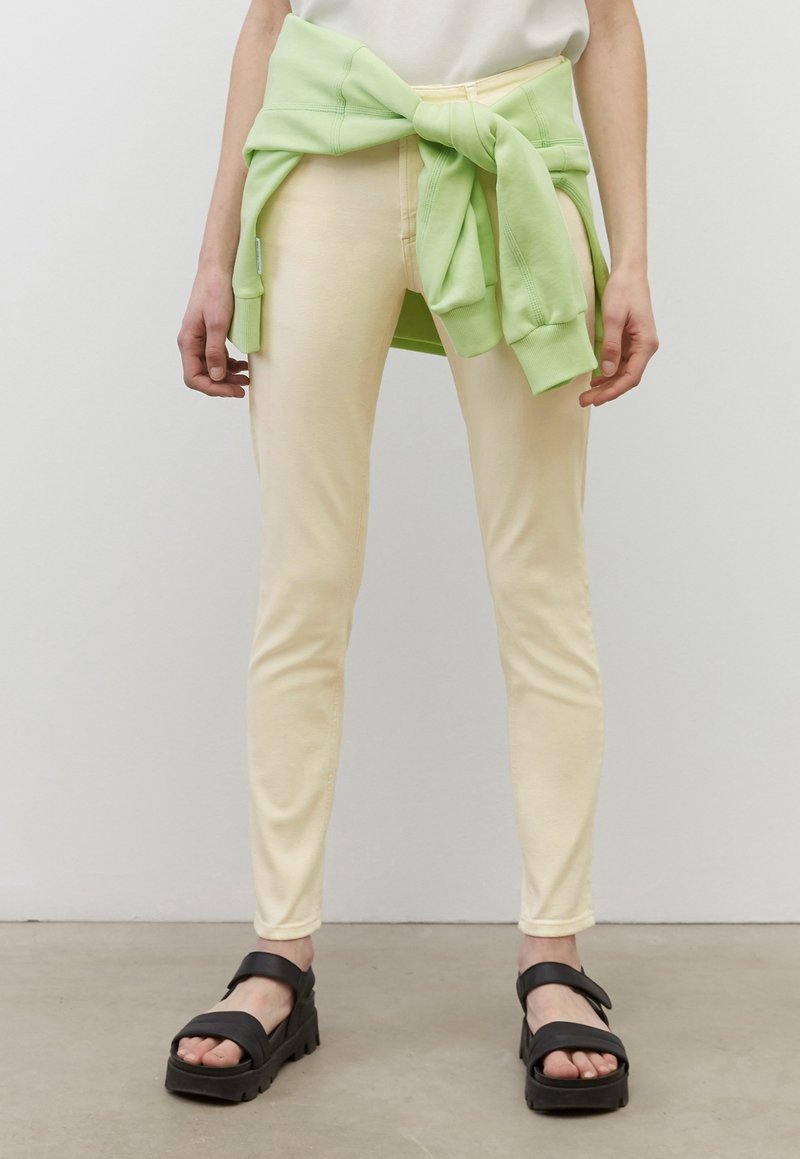Pantalons jaunes évasés en tissu doux, associés à un sweat-shirt vert clair noué autour de la taille ; sandales à plateforme noires.