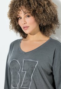 Graues Langarmshirt mit einer großen gestickten Zahl "87" und dekorativen Mustern, V-Ausschnitt und weichem Baumwollstoff.