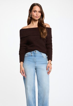 Kvinde iført en mørk off-shoulder sweater, lyseblå høj-taljede jeans og gyldne hoop-øreringe, stående mod en hvid baggrund.
