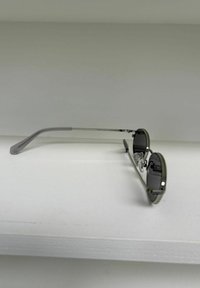 Lunettes de soleil rondes à monture métallique fine avec des verres foncés reposant sur une surface blanche devant un fond gris uni.