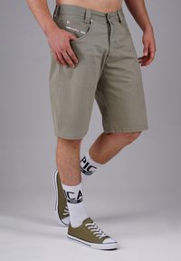 Khaki-Shorts mit geradem Schnitt, Fronttaschen und Kupferbeschlägen, getragen mit olivgrünen Canvas-Sneakern und weißen Socken mit schwarzem Text.