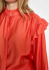 Blusa in satin rosso con colletto e maniche arricciate, dotata di una chiusura con bottoni e una textura liscia. Finitura leggermente lucida.