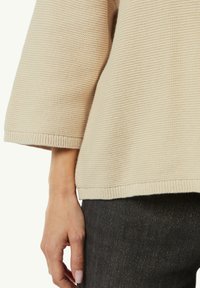 Beige strukturierter Pullover mit Dreiviertelärmel, ausgestattet mit einem gerippten Saum und Manschetten, kombiniert mit dunklen Denim-Hosen.