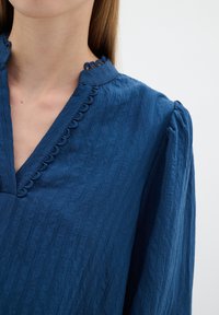 Kék, textúrált blúz V nyakkivágással, fodros díszítéssel és puffos ujjal. Az anyaga enyhe gyűrött hatású.