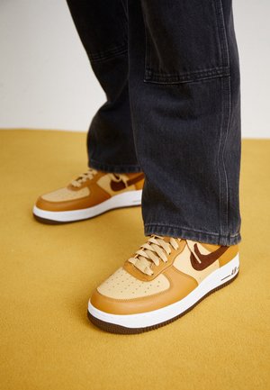 Personne portant des baskets Nike Air Force 1 beige et marron et un pantalon en jean noir ample, debout sur un sol jaune moutarde.