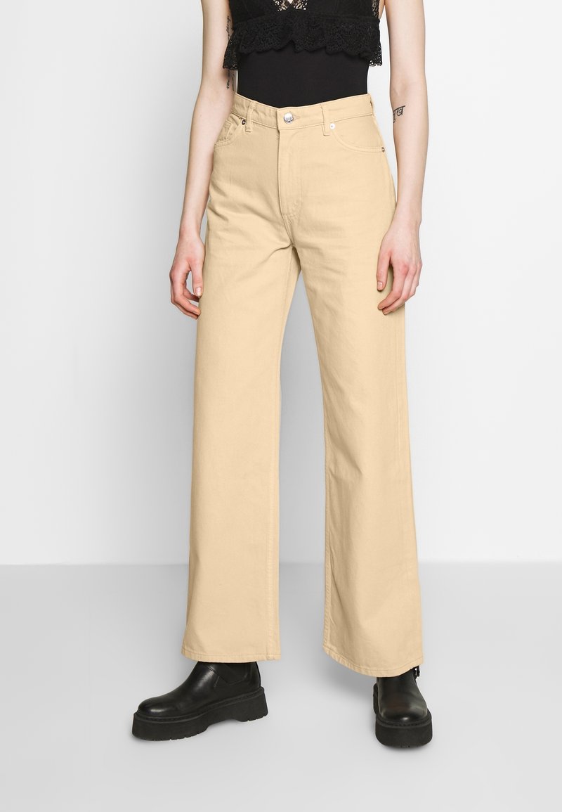 Pantalones de pierna ancha beige hechos de algodón, con una cintura alta y dos bolsillos frontales, combinados con botas negras gruesas.