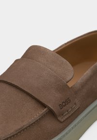 Mocassin en daim marron avec un détail de bride et le logo "BOSS" embossé près de la couture au-dessus de la semelle en caoutchouc claire.