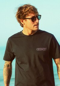 T-shirt in cotone nero con logo viola "OXBOW", colletto rotondo e maniche corte. Il modello indossa occhiali da sole neri; sullo sfondo si vede l'oceano.