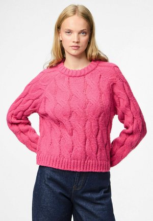 Blonde vrouw in een felroze kabelgebreide trui en donkerblauwe jeans, staande met haar handen op haar heupen tegen een witte achtergrond.