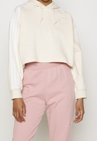 Oříznutá béžová mikina s kapucí a stahovací šňůrkou, s bílými pruhy po stranách a vyšitým logem. Kombinována s měkkými růžovými tepláky.