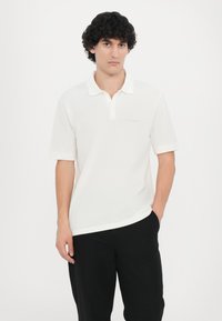Biela polo tričko s golierom, krátkymi rukávmi a vyšitým logom na ľavej strane. V kombinácii s čiernymi nohavicami, model stojí na bielom pozadí.