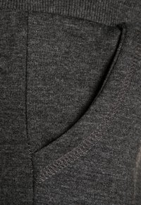 Grijze stof met een close-up weergave van een zak, met een gladde textuur en zichtbare stiksels langs de randen. Subtiele kleurvariaties aanwezig.