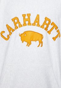 Weißer Stoff mit gelber, gestickter Schrift "CARHARTT", die sich oberhalb einer gelben, gestickten Silhouette eines Bisons in der Mitte darunter wölbt.
