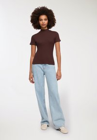 Brązowy dopasowany top z krótkim rękawem i wysokim dekoltem, połączony z jasnoniebieskimi szerokimi spodniami dżinsowymi i beżowymi trampkami, na jednolitym tle.