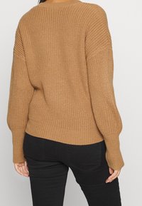 Hnědý pletený svetr s žebrovanými manžetami a lemem, s volným střihem a strukturovaným vzorem. Pohled zezadu na jednoduchém pozadí.