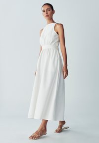 Weißes ärmelloses Kleid mit geraffter Taille, fließendem Stoff und knöchellangem Saum. Getragen mit beigen Riemchensandalen. Einfaches, modernes Design.