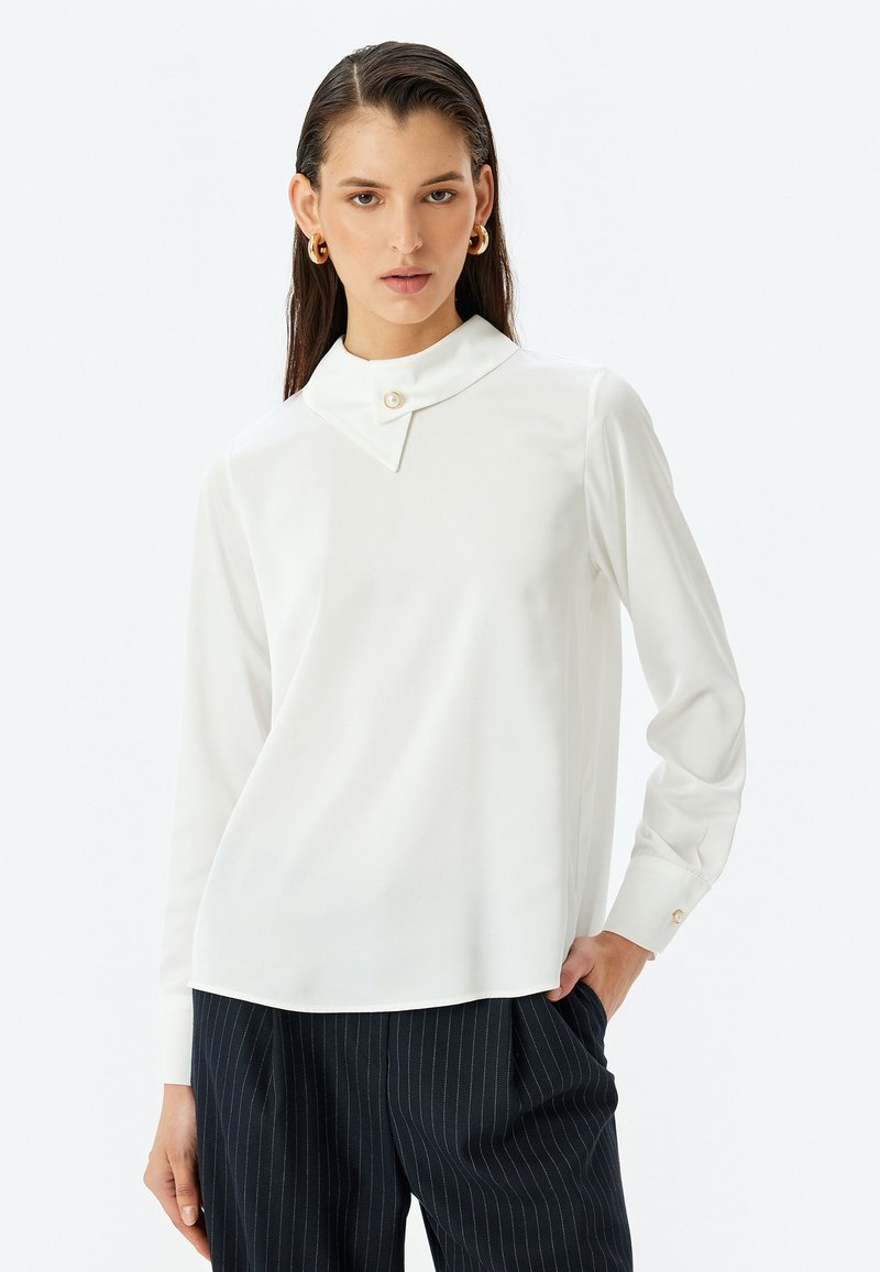 Weiße Bluse mit langen Ärmeln, spitzen Kragen und Knopfdetaillierung, mit glatter Textur. Getragen mit gestreiften marineblauen Hosen.