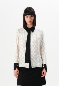 White blouse with black geometric pattern, black collar, and tie. Long sleeves with black cuffs and gold button accents. Worn over a black skirt.