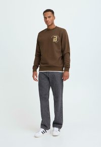 Bruine sweatshirt met een ronde halslijn en een gouden logo-accent, gecombineerd met donkergrijze jeans en witte sneakers met zwarte strepen.