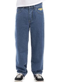 Modri denim hlače imajo sproščen kroj z vidnimi šivi in šivanjem. Sprednji žepi vključujejo logo na desni strani. Nosi se črne superge.