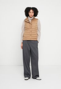 Tan brūns pūkains vesti ar augstu apkakli, priekšējām pogām un krokotu dizainu, valkāts virs gaiša džempera. Saskaņots ar pelēkamiem platām biksēm un apaviem.