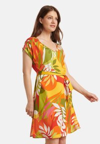 Bloemenjurk met een V-hals, korte mouwen en een strik in de taille. De stof toont levendige, meerkleurige bladerenpatronen op een gele achtergrond.