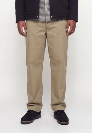 Homme portant un pantalon beige droit, des chaussures noires et une veste noire par-dessus une chemise à carreaux, debout devant un fond clair uni.