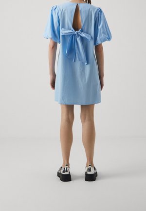 Femme debout, vue de dos, portant une robe courte bleu clair à manches bouffantes et un grand nœud noué dans le dos ouvert, chaussures à plateforme argentées.