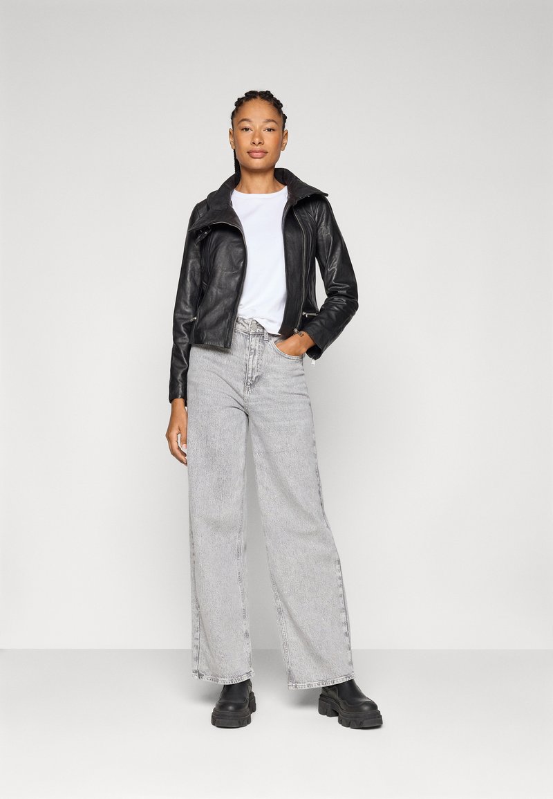 Frau mit geflochtenem Haar, die eine schwarze Lederjacke, ein weißes T-Shirt, helle graue weit geschnittene Jeans und schwarze klobige Stiefel trägt und vor einem neutralen Hintergrund steht.